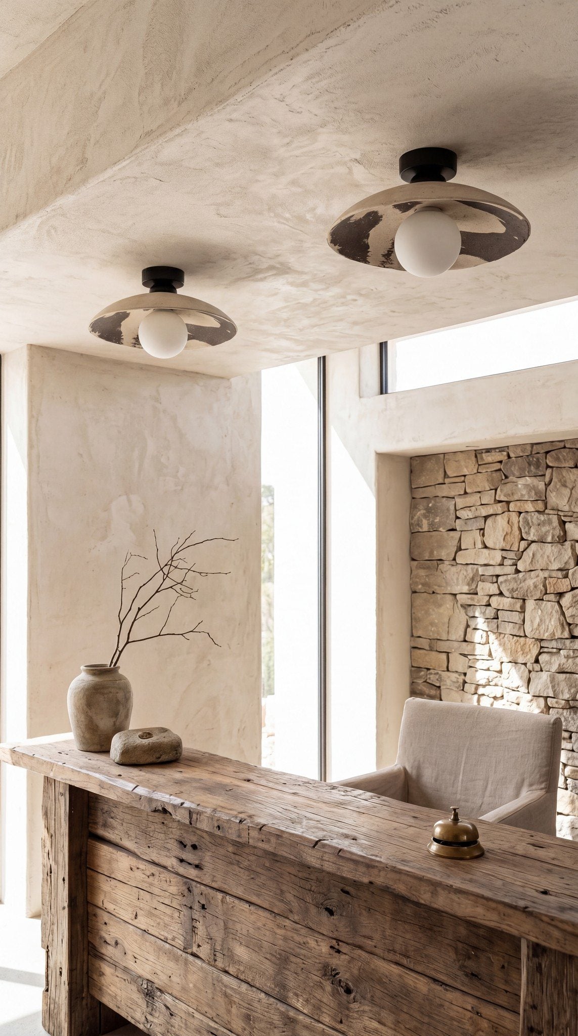 Two Nara ceramic flush mount ceiling lights above a reclaimed wood hotel reception desk with stone wall